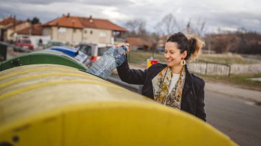 Bir yetişkin kadın, plastik atık ve sarı poşetlerle geri dönüşüm kutularının önünde duruyor ve sıfır atık eko kavramını yeniden kullanmak için şişe fırlatıyor.