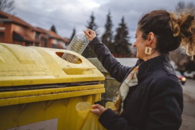 Bir yetişkin kadın, plastik atık ve sarı poşetlerle geri dönüşüm kutularının önünde duruyor ve sıfır atık eko kavramını yeniden kullanmak için şişe fırlatıyor.