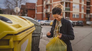 Bir yetişkin kadın, plastik atık ve sarı poşetlerle geri dönüşüm kutularının önünde duruyor ve sıfır atık eko kavramını yeniden kullanmak için şişe fırlatıyor.