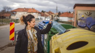 Bir yetişkin kadın, plastik atık ve sarı poşetlerle geri dönüşüm kutularının önünde duruyor ve sıfır atık eko kavramını yeniden kullanmak için şişe fırlatıyor.