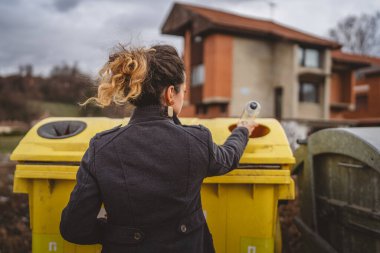 Bir yetişkin kadın, plastik atık ve sarı poşetlerle geri dönüşüm kutularının önünde duruyor ve sıfır atık eko kavramını yeniden kullanmak için şişe fırlatıyor.
