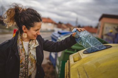 Bir yetişkin kadın, plastik atık ve sarı poşetlerle geri dönüşüm kutularının önünde duruyor ve sıfır atık eko kavramını yeniden kullanmak için şişe fırlatıyor.