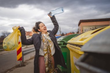 Bir yetişkin kadın, plastik atık ve sarı poşetlerle geri dönüşüm kutularının önünde duruyor ve sıfır atık eko kavramını yeniden kullanmak için şişe fırlatıyor.