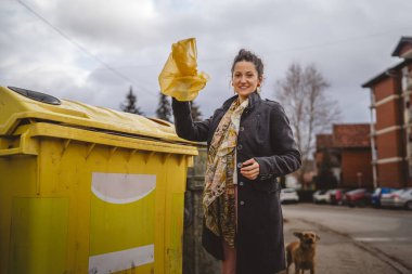 Bir yetişkin kadın, plastik atık ve sarı poşetlerle geri dönüşüm kutularının önünde duruyor ve sıfır atık eko kavramını yeniden kullanmak için şişe fırlatıyor.
