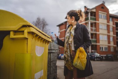 Bir yetişkin kadın, plastik atık ve sarı poşetlerle geri dönüşüm kutularının önünde duruyor ve sıfır atık eko kavramını yeniden kullanmak için şişe fırlatıyor.