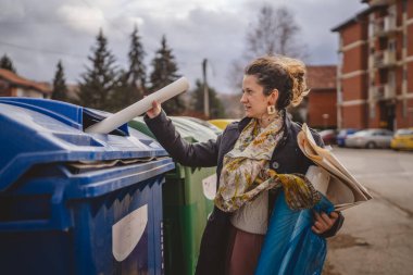 Bir yetişkin kadın kağıt atık ve mavi poşetlerle geri dönüşüm kaplarının önünde duruyor ve sıfır atık eko kavramını yeniden kullanmak için atıyorlar.