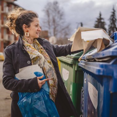 Bir yetişkin kadın kağıt atık ve mavi poşetlerle geri dönüşüm kaplarının önünde duruyor ve sıfır atık eko kavramını yeniden kullanmak için atıyorlar.