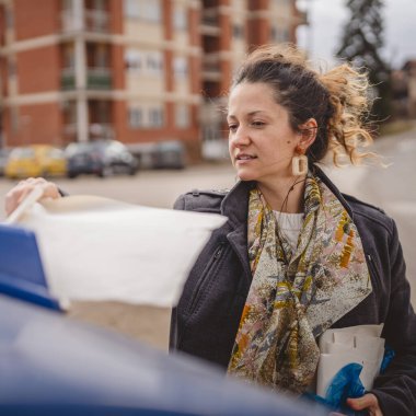 Bir yetişkin kadın kağıt atık ve mavi poşetlerle geri dönüşüm kaplarının önünde duruyor ve sıfır atık eko kavramını yeniden kullanmak için atıyorlar.