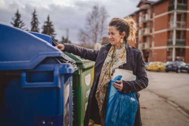 Bir yetişkin kadın kağıt atık ve mavi poşetlerle geri dönüşüm kaplarının önünde duruyor ve sıfır atık eko kavramını yeniden kullanmak için atıyorlar.