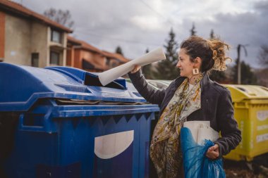 Bir yetişkin kadın kağıt atık ve mavi poşetlerle geri dönüşüm kaplarının önünde duruyor ve sıfır atık eko kavramını yeniden kullanmak için atıyorlar.