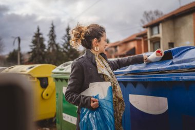 Bir yetişkin kadın kağıt atık ve mavi poşetlerle geri dönüşüm kaplarının önünde duruyor ve sıfır atık eko kavramını yeniden kullanmak için atıyorlar.