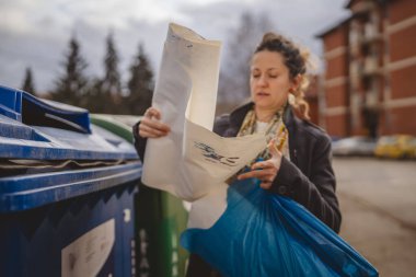 Bir yetişkin kadın kağıt atık ve mavi poşetlerle geri dönüşüm kaplarının önünde duruyor ve sıfır atık eko kavramını yeniden kullanmak için atıyorlar.