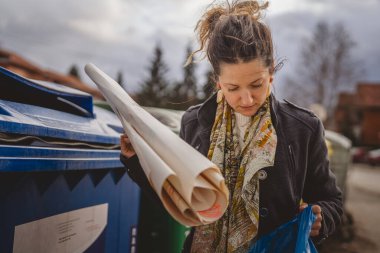 Bir yetişkin kadın kağıt atık ve mavi poşetlerle geri dönüşüm kaplarının önünde duruyor ve sıfır atık eko kavramını yeniden kullanmak için atıyorlar.