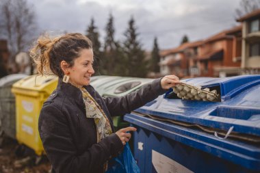 Bir yetişkin kadın kağıt atık ve mavi poşetlerle geri dönüşüm kaplarının önünde duruyor ve sıfır atık eko kavramını yeniden kullanmak için atıyorlar.