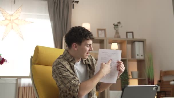 年轻男子高加索少年坐在家里用数码平板电脑展示论文书信或奖学金合同考试结果在家里打一个视频电话开心地笑着 — 图库视频影像