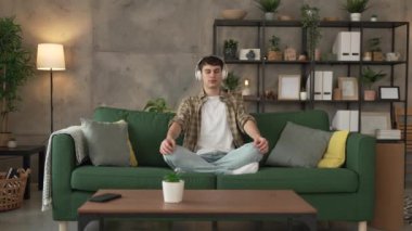 Genç bir beyaz erkek, internet rehberli meditasyon farkındalık yogası için kulaklık kullanıyor. Evde gözleri kapalı, gerçek insanlar kişisel bakım tezahürü konsepti Z jenerasyonunu taklit ediyor.