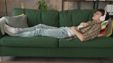 Genç bir beyaz erkek, internet rehberli meditasyon farkındalık yogası için kulaklık kullanıyor. Evde gözleri kapalı, gerçek insanlar kişisel bakım tezahürü konsepti Z jenerasyonunu taklit ediyor.