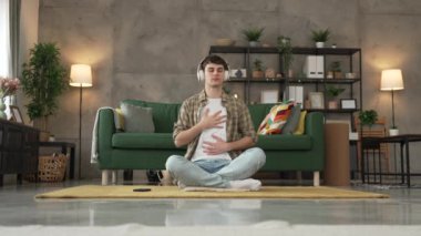 Genç bir beyaz erkek, internet rehberli meditasyon farkındalık yogası için kulaklık kullanıyor. Evde gözleri kapalı, gerçek insanlar kişisel bakım tezahürü konsepti Z jenerasyonunu taklit ediyor.