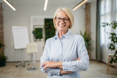 Ofiste gözlüklü, olgun, beyaz bir kadının portresi. Mutlu bir gülümseme. Kendine güvenen fotoğraf makinesine bakıyor.