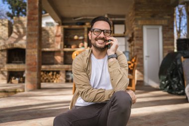 Sakallı ve gözlüklü yetişkin bir adam güneşli bir günde dışarıda oturur mutlu bir şekilde gülümser cep telefonunu kullanarak konuşma yeri fotokopisi çektirir gerçek insanlar gibi.