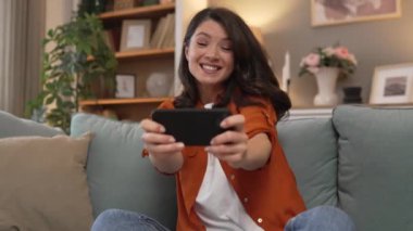 Yetişkin beyaz bir kadın evinde mutlu bir şekilde oturup video oyunları oynayıp eğleniyor cep telefonu kullanıyor ve internet üzerinden eğleniyor.