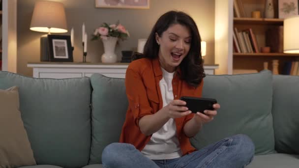 一人の大人の白人女性は 楽しい携帯電話の使用スマートフォンを保持している楽しいビデオゲームレジャー活動を家に座って オンラインで楽しいです — ストック動画