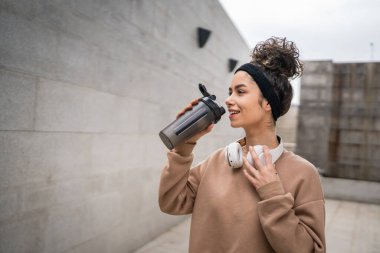Kulaklıklı bir genç kadın eğitim için hazırlanıyor. Su içmek için destek şişesi tutuyor. Açık havada güzel beyaz kadın nesli.