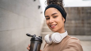 Kulaklıklı bir genç kadın eğitim için hazırlanıyor. Su içmek için destek şişesi tutuyor. Açık havada su içmek için güzel beyaz kadın selfie sağlıklı yaşam tarzı UGC Kullanıcı tarafından oluşturulmuş.