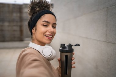 Kulaklıklı bir genç kadın eğitim için hazırlanıyor. Su içmek için destek şişesi tutuyor. Açık havada su içmek için güzel beyaz kadın selfie sağlıklı yaşam tarzı UGC Kullanıcı tarafından oluşturulmuş.