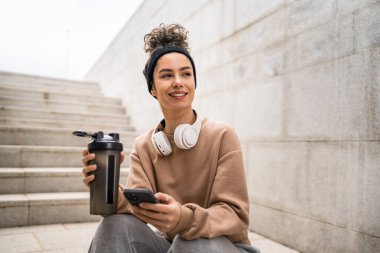 Kulaklık takan bir genç kadın destek şişesi ve akıllı telefon, spor, Kafkasyalı bir kadın Z jenerasyonu sağlıklı yaşam tarzı konseptini kopyalamak için fren yapıyor.