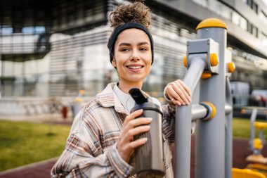 Kulaklıklı bir genç kadın eğitim için hazırlanıyor. Su içmek için destek şişesi tutuyor. Açık havada güzel beyaz kadın nesli.