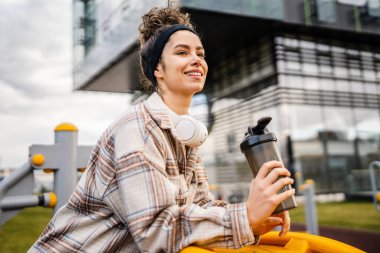 Kulaklıklı bir genç kadın eğitim için hazırlanıyor. Su içmek için destek şişesi tutuyor. Açık havada güzel beyaz kadın nesli.