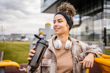 Kulaklıklı bir genç kadın eğitim için hazırlanıyor. Su içmek için destek şişesi tutuyor. Açık havada güzel beyaz kadın nesli.