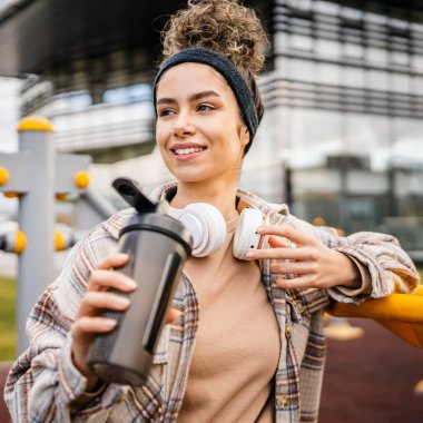 Kulaklıklı bir genç kadın eğitim için hazırlanıyor. Su içmek için destek şişesi tutuyor. Açık havada güzel beyaz kadın nesli.