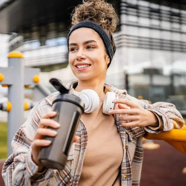 Kulaklıklı bir genç kadın eğitim için hazırlanıyor. Su içmek için destek şişesi tutuyor. Açık havada güzel beyaz kadın nesli.