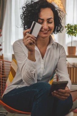 Evde mutlu gülümseyen yetişkin bir kadın banka kredi kartı ve cep telefonu online alışveriş e-bankacılığı konseptini tutuyor. Gerçek insanlar uzay e-ticaretini kopyalayıp rezervasyon yaptırıyor.