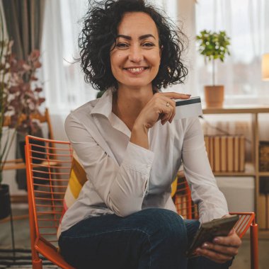 Evde mutlu gülümseyen yetişkin bir kadın banka kredi kartı ve cep telefonu online alışveriş e-bankacılığı konseptini tutuyor. Gerçek insanlar uzay e-ticaretini kopyalayıp rezervasyon yaptırıyor.