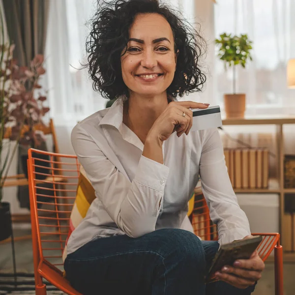 Evde mutlu gülümseyen yetişkin bir kadın banka kredi kartı ve cep telefonu online alışveriş e-bankacılığı konseptini tutuyor. Gerçek insanlar uzay e-ticaretini kopyalayıp rezervasyon yaptırıyor.