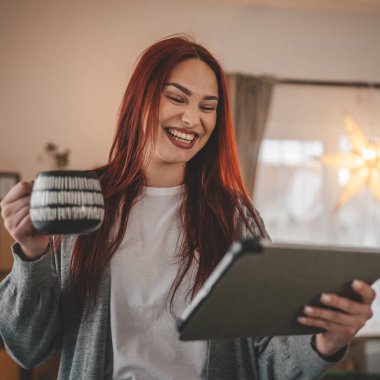 Kadın elinde dijital tablet ve kahveyle pencerede dikiliyor.
