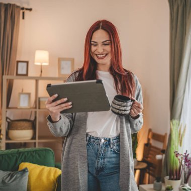 Kadın elinde dijital tablet ve kahveyle pencerede dikiliyor.