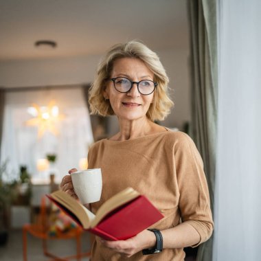 Yetişkin bir beyaz kadın evde kitap okuyor ve gözlük takıyor.