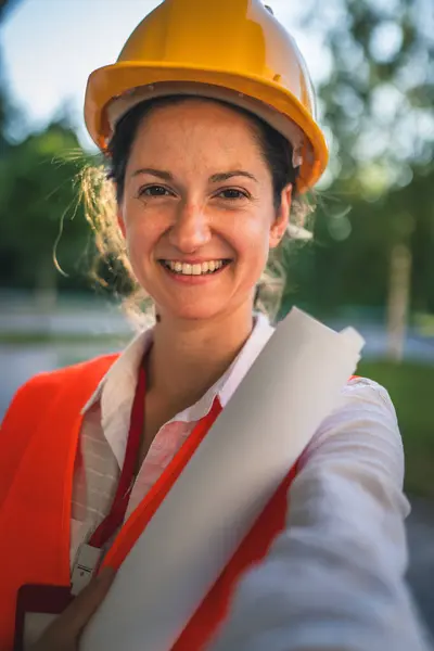 architect woman female construction engineer stand outdoors wear protective helmet and west in front of modern building wall self portrait selfie UGC user generated content