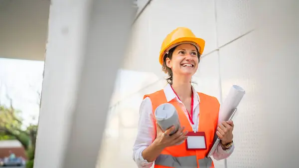 architect woman female construction engineer stand outdoors wear protective helmet and west in front of modern building wall
