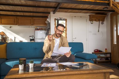 Evden çalışan yetişkin bir beyaz adam kanepeye oturur telefon görüşmesi yapar. Bu arada evraklar, iş raporları ya da belgeler elinde olur.