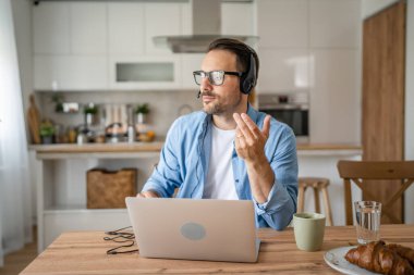 Kafkasyalı bir erkek, kafasında kulaklıkla evde çalışıyor. Bilgisayardaki müşteri desteğiyle ciddi düşünüyor.