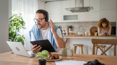 Kafkasyalı bir erkek, evinde kulaklıkla bilgisayar başında çalışıyor. Müşteri destekliyor. Karısının arka planda olduğunu düşünüyor.