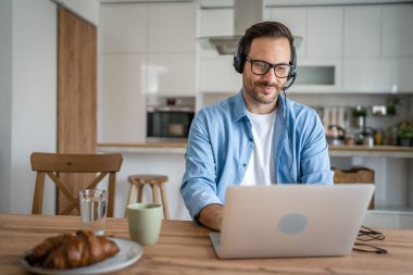 Kafkasyalı bir erkek bilgisayar başında kulaklıkla evde çalışıyor. Müşteri mutlu bir gülümsemeyi destekliyor.