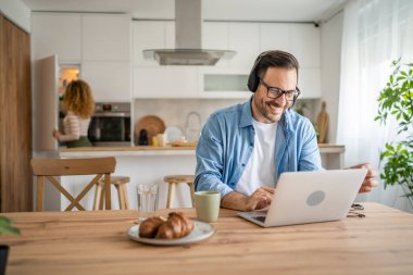 Kafkasyalı bir erkek bilgisayar başında kulaklıkla evden çalışıyor. Müşteri mutlu bir gülümsemeyi destekliyor. Karısının arkada durduğundan emin.