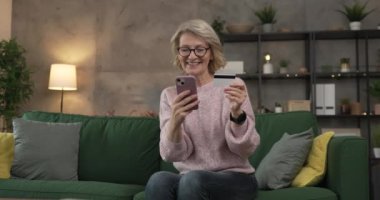 Olgun beyaz kadın kredi kartını evde online alışveriş için tutuyor.