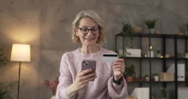 Olgun beyaz kadın kredi kartını evde online alışveriş için tutuyor.